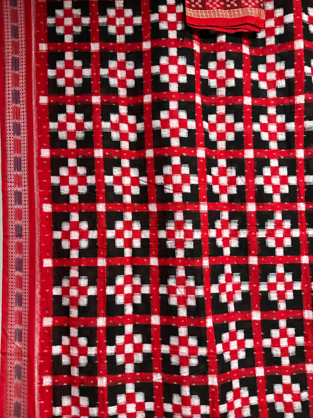 A close-up of a red, black, and white patterned fabric, likely a traditional Indian saree or textile, featuring a geometric ikat design with a red border.